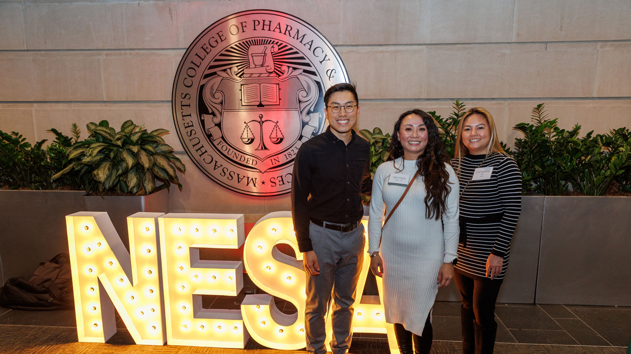 Three people stand in front of the MCPHS seal and NESA lighted letters.